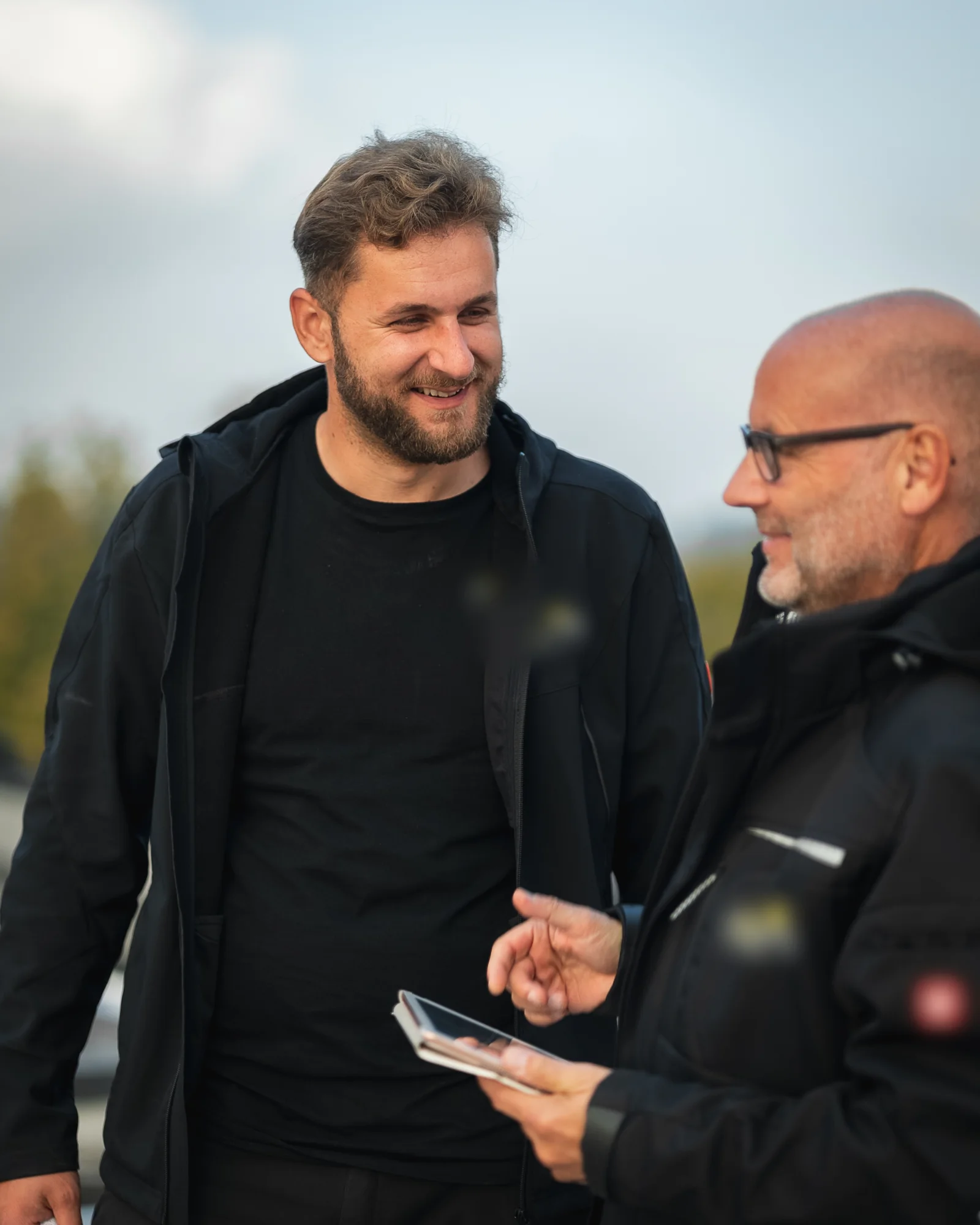 Two site managers in black jackets talk outdoors; the older one holds a tablet.