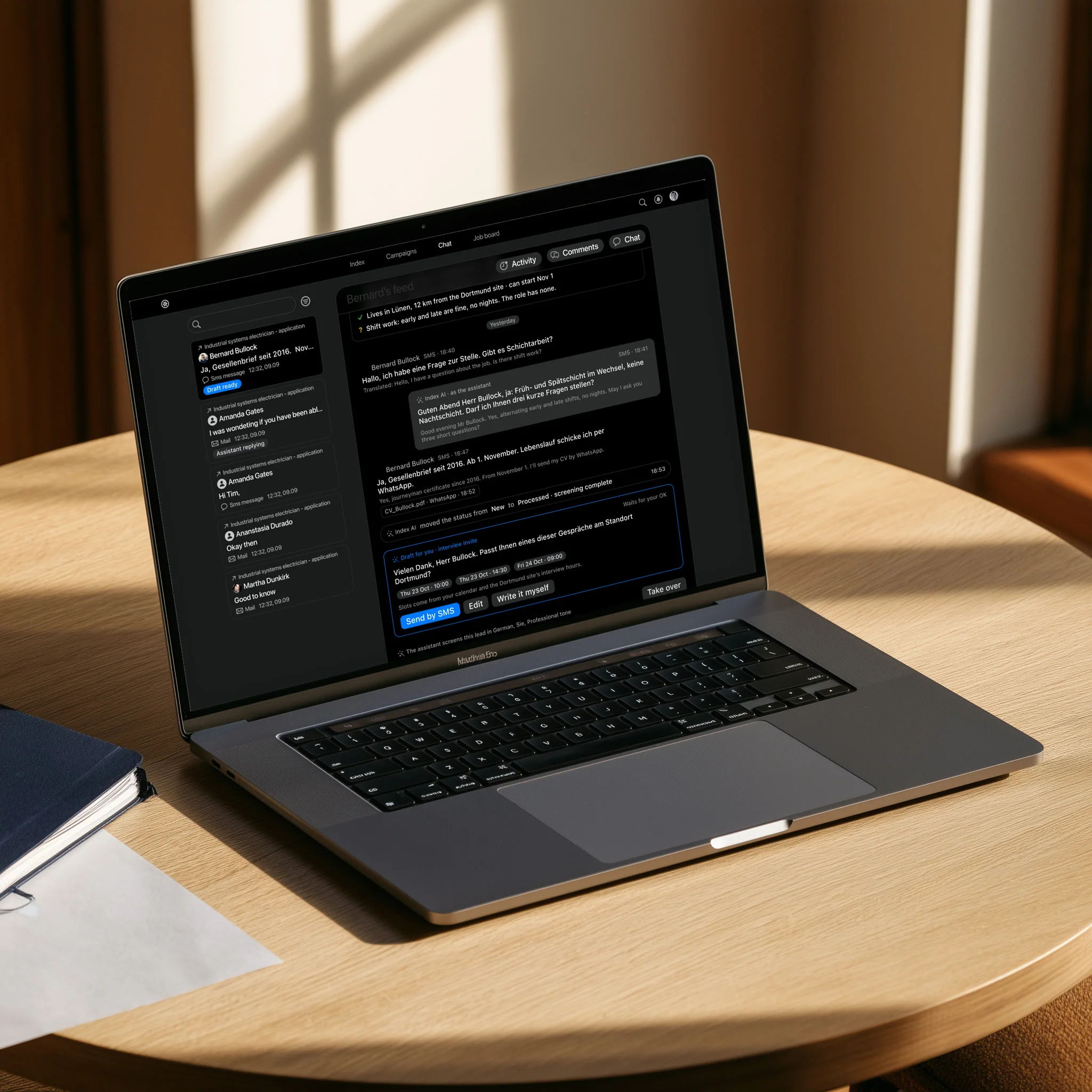 A MacBook Pro on a round oak table shows the Index chat with an applicant, where the assistant offers interview slots.