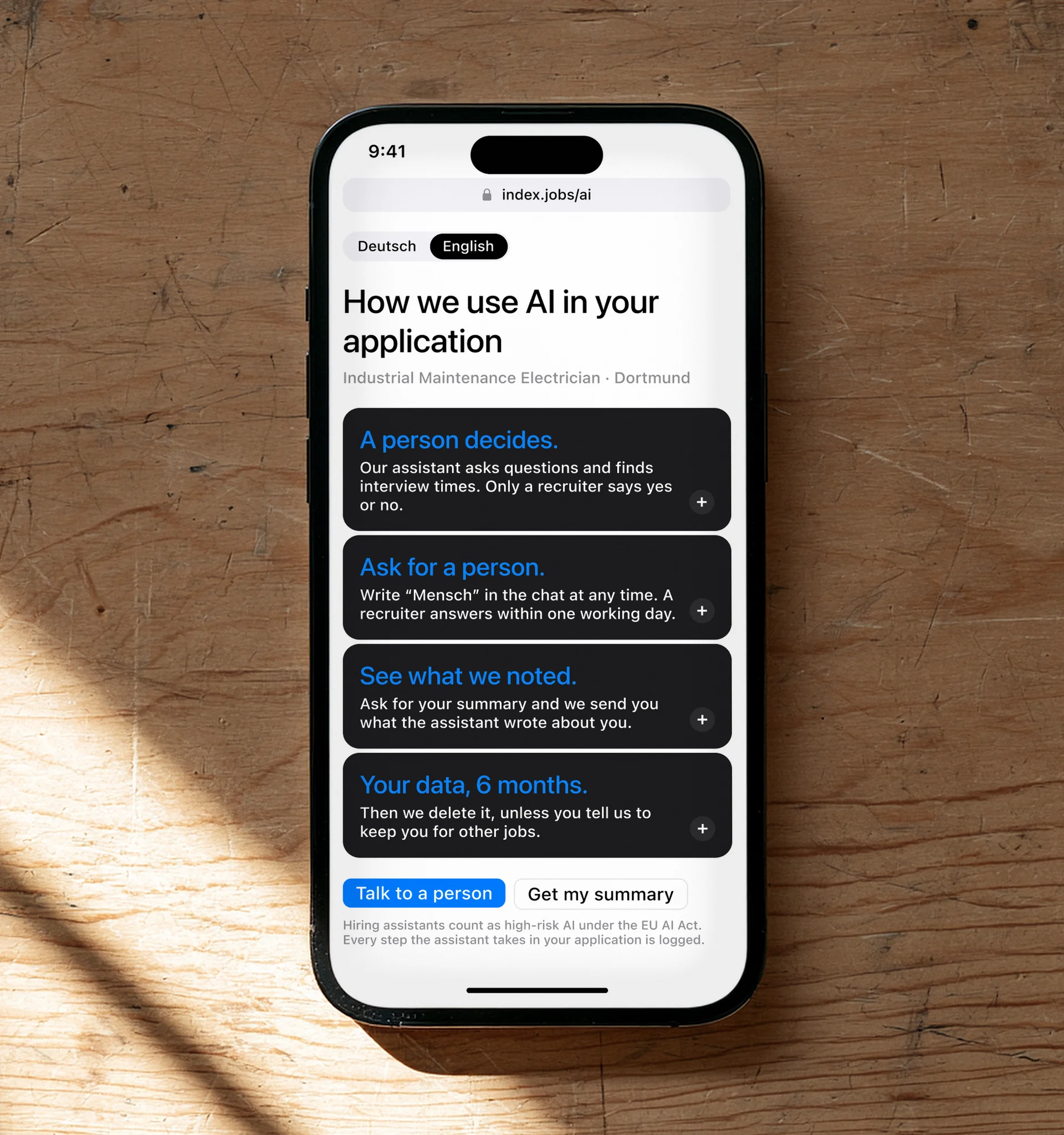 An iPhone on a plywood workbench showing the Index page How we use AI in your application: a person decides, ask for a person, see what we noted, your data for six months.