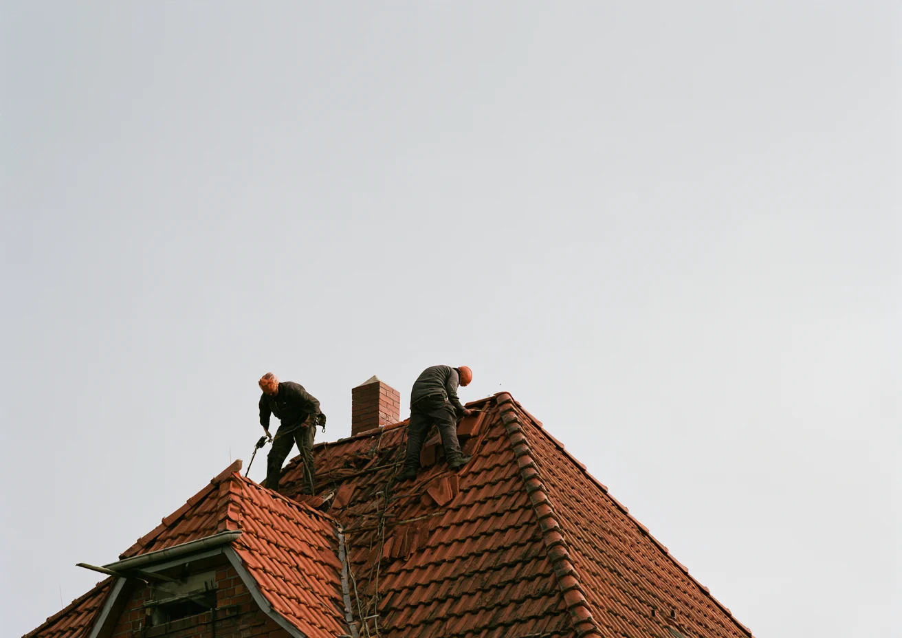Two roofers lay tiles on a steep red roof.