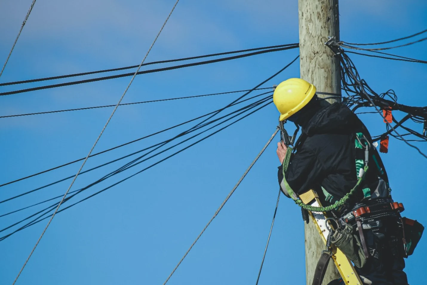 An electrician in a yellow helmet works at the top of a wooden pole against a blue sky.