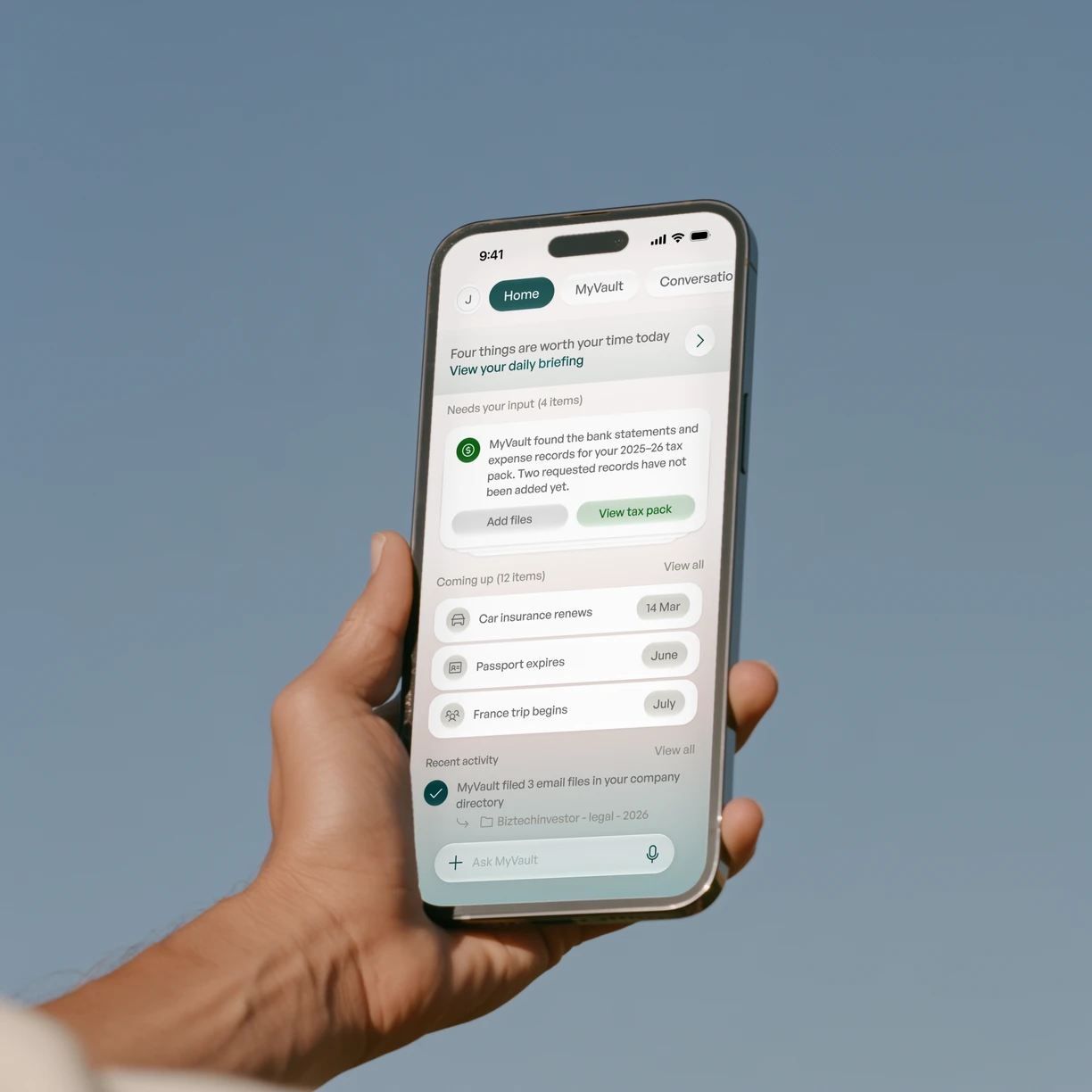 A hand holds an iPhone up against a blue sky, open on MyVault's Home: a daily briefing banner, a tax pack that needs two files, and what's coming up.