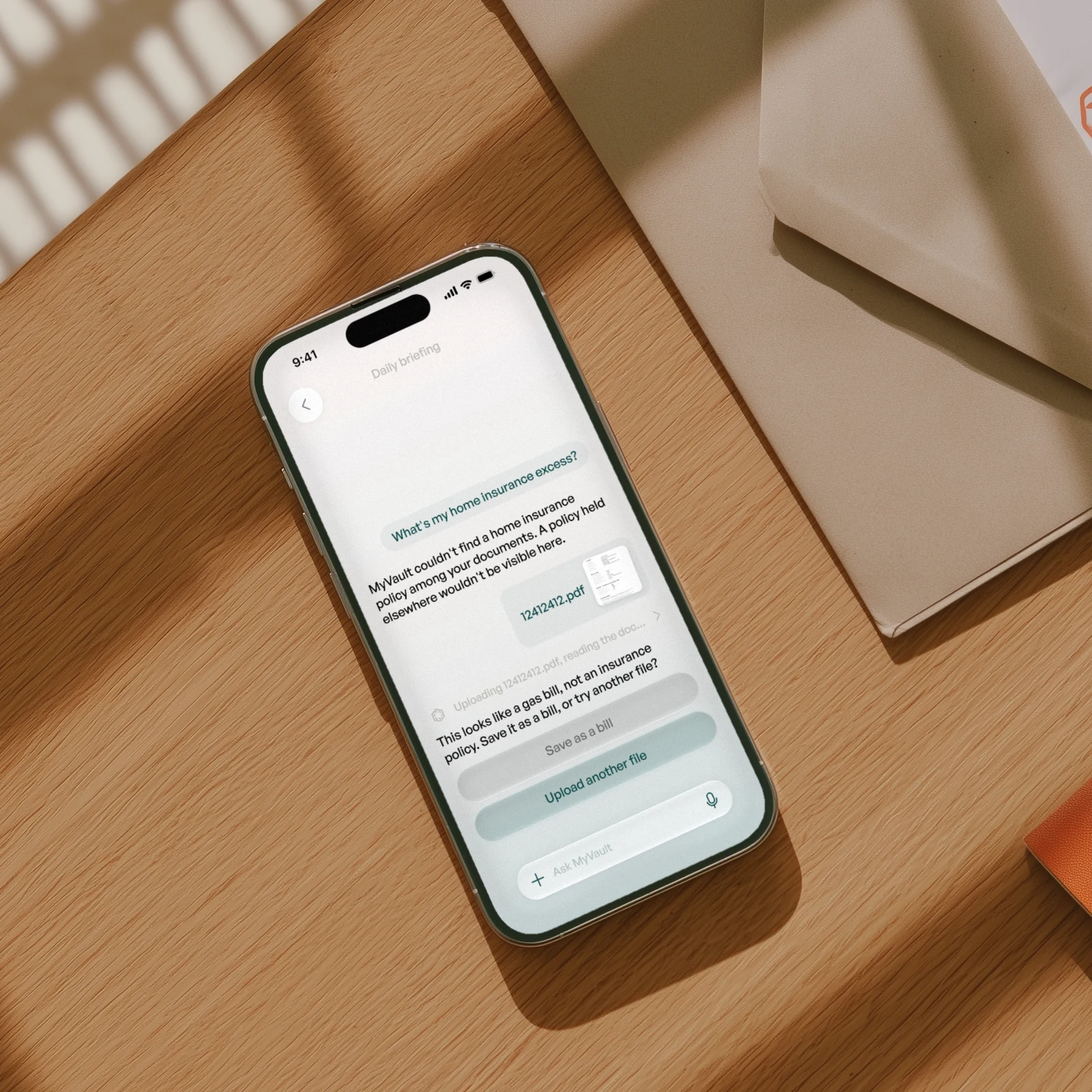 An iPhone on an oak desk beside a kraft envelope, open on MyVault: asked for the home insurance excess, it says it can't find a policy, reads the uploaded PDF, and offers to save it as a gas bill instead