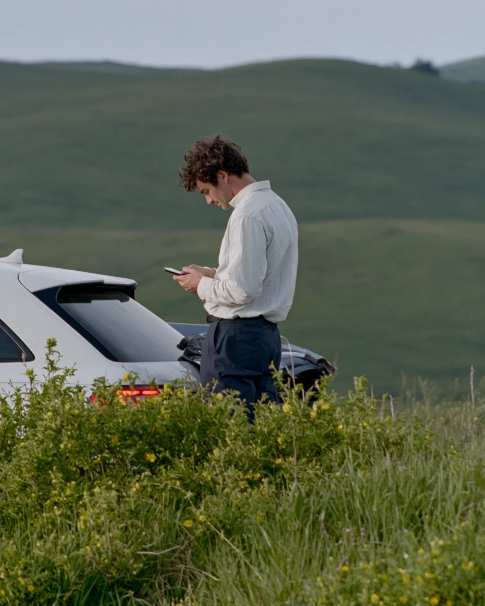 A man in a white shirt checks his phone beside his car on a green hillside road